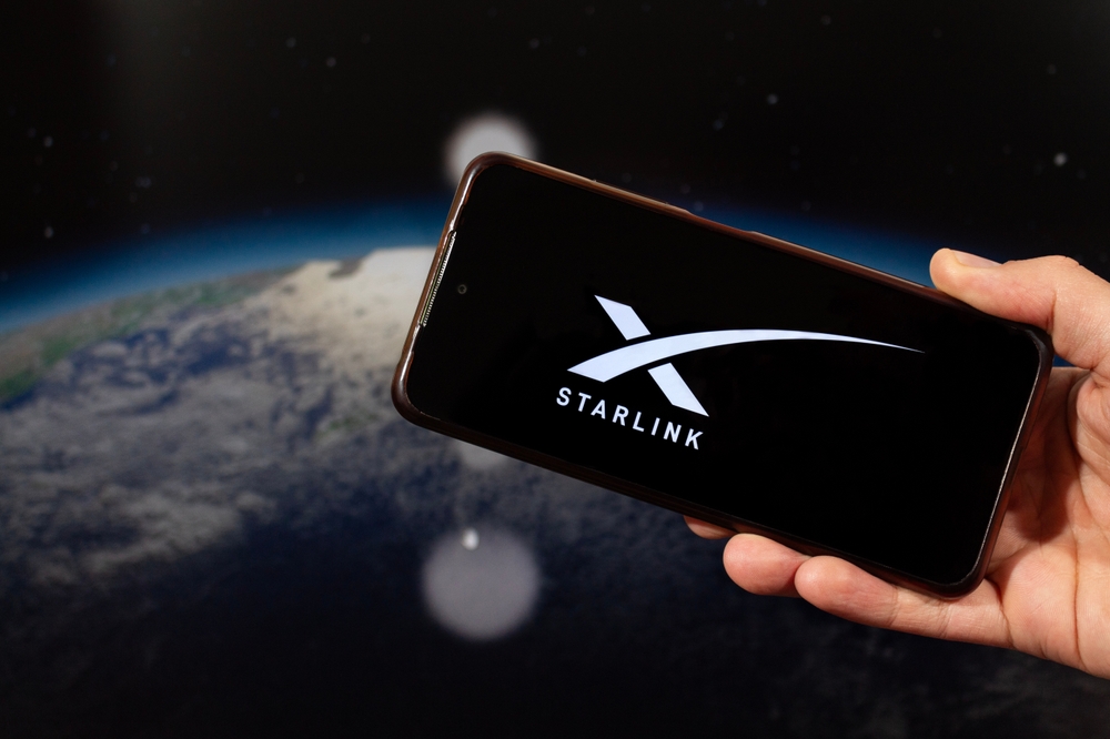 A hand holding a phone displaying the Starlink logo, with the curve of the Earth visible behind it.