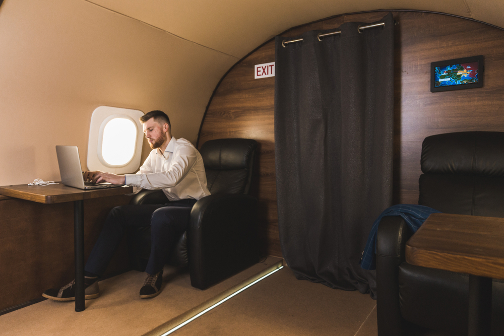 A businessman working on a laptop at a cabin table aboard a private jet, seated beside the window.